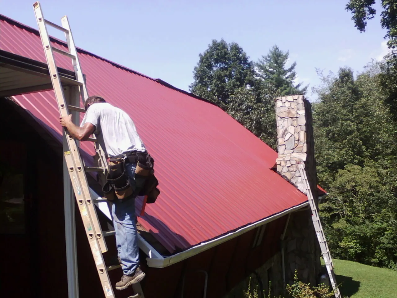 Roofer on a ladder beside a red metal roof and stone chimney — Soffit & Fascia Repair by Roofing Brothers in Quail Valley